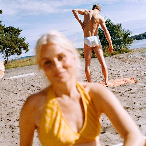 Kvinna på strand i förgrunden med man i badkläder bakom
